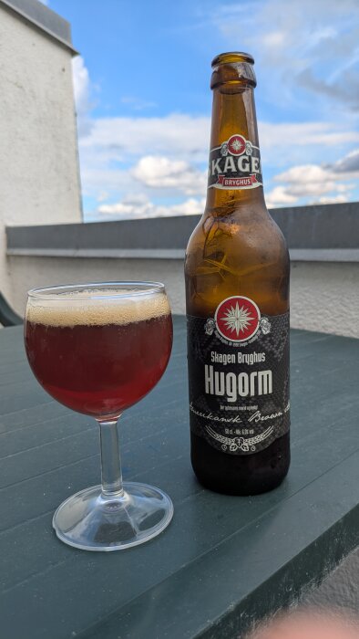 Flaska och glas med brun ale, märkt "Skagen Bryghus Hugorm", står på ett bord utomhus under en blå himmel.