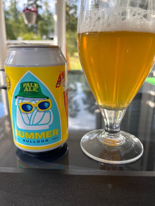 En burk och ett glas med Summer Bulldog Pale Ale på ett bord, omgiven av grönska i bakgrunden.