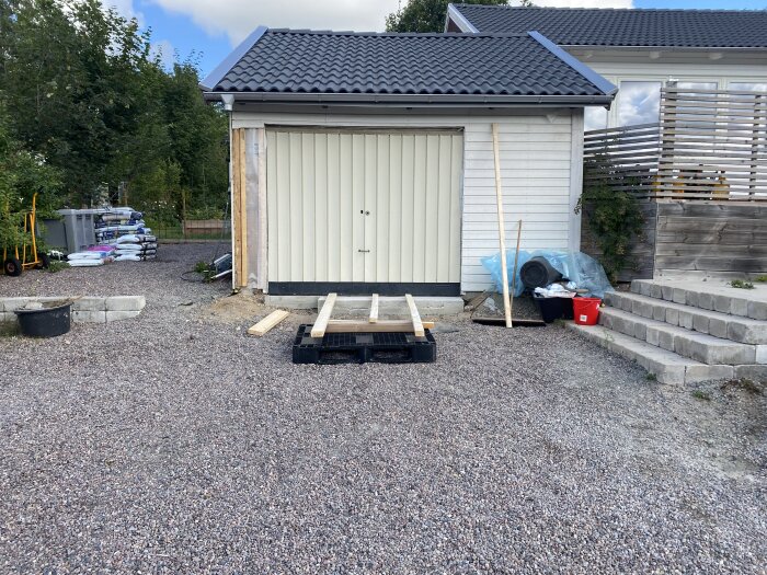 Garage med bytt port, omgivande grusplan och byggmaterial på marken, inklusive träplankor och betongblock.