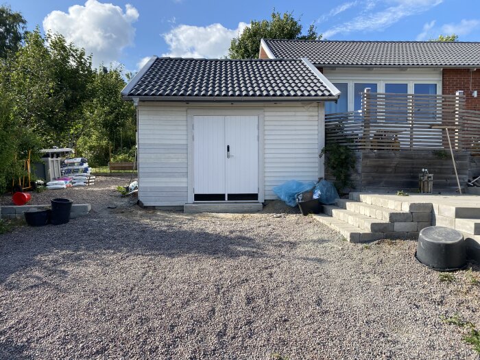 Vit garagebyggnad med ny dubbeldörr, flankerad av trappa i sten och grusförgård; solig dag med blå himmel och träd i bakgrunden.