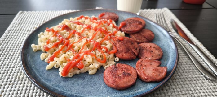 En tallrik med makaroner toppade med ketchup och bredvid skivor av stekt korv, på en bordstablett med bestick och ett glas mjölk i bakgrunden.