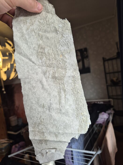 En hand håller i en reva av gammal, vit väv som verkar vara slipad, med slitet och ojämnt ytskikt. I bakgrunden syns inomhusdetaljer.