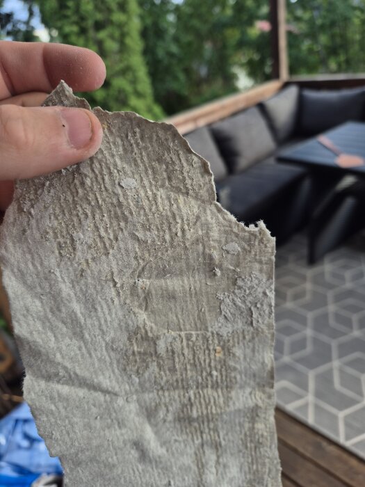 En hand håller en bit grå, skrovlig väv, potentiellt asbest eller glasfiber, med en uteplats med soffa och bord i bakgrunden.