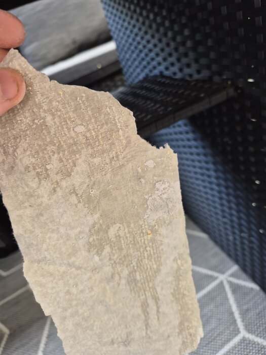 Bit av gammal väv med struktur, hålls framför en flätad yta; osäkert material, eventuell asbest eller glasfiber från ett 50-talshus.