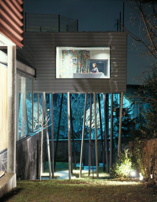 Modern byggnad svävande på höga pelare, med stort, belyst fönster där en person syns inomhus, omgiven av träd och belysning i en nattmiljö.