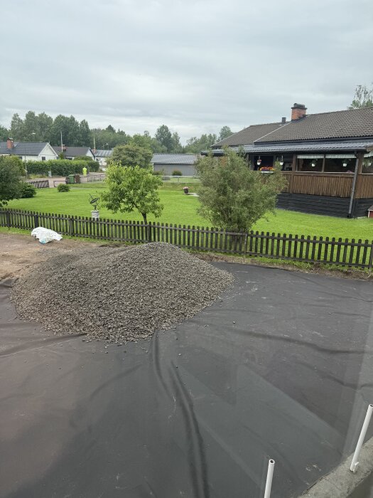 Hög med grus på förberedd uppfart med svart markduk, omgiven av trädgård och staket. En del av ett projekt för att dränera och anlägga uppfart.