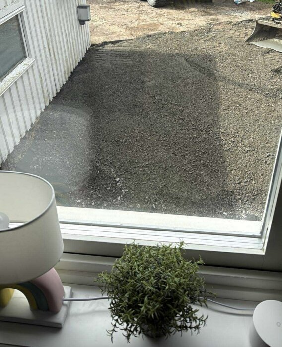 Vy från ett fönster mot en ny grusuppfart och ett hörn av ett hus. En krukväxt och lampa är synliga på fönsterbrädan.