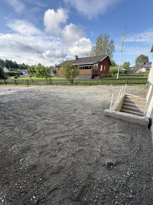 Nygrusad uppfart med bärlager i fokus, hus och trädgård i bakgrunden, samt trappa med cementarmering synlig längst ner.