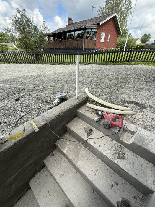 Betongtrappa med borrmaskin och dräneringsslang i arbete, grusuppfart i bakgrunden, hus och staket syns längre bort.
