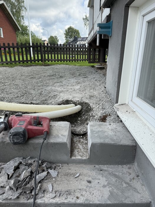 Vit dräneringsslang placerad i urgröpning i cementtrappa, omgiven av grus och krossad betong, med en röd kapmaskin i förgrunden.