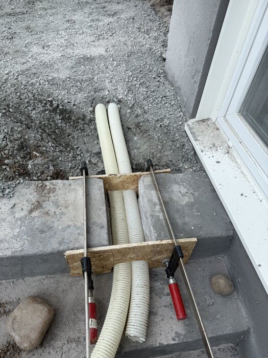 Två dräneringsslangar inplacerade vid källartrappa, inkapslade i cement med form av OSB-skivor, omgivna av grus och stenklampar.