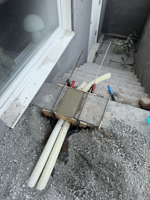 Dräneringsslangar i källartrappa, inbäddade i cement med OSB-form, omgivna av grus och verktyg, efter renoveringsarbete.