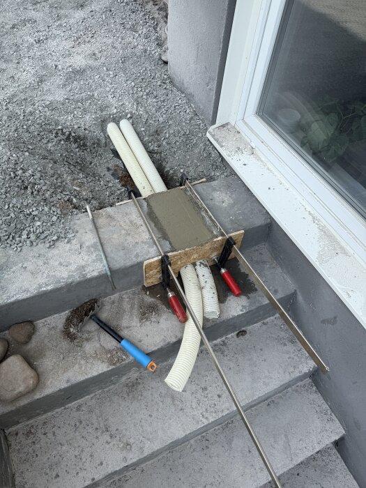 Dräneringsslangar installeras i källartrappa med cement och OSB-skivor; grus syns i bakgrunden. Verktyg används för installationen.