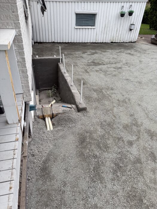 Bärlager av grus på uppfart; dräneringsslang dold vid källartrappa med färsk cementgjutning; vit byggnad och planteringskärl i bakgrunden.