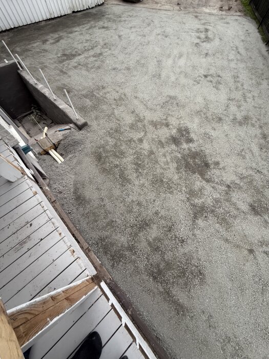 Nygrusad uppfart med synlig dräneringsslang inbäddad i cement vid sidan av en källartrappa, omgiven av vita stängseldelar.