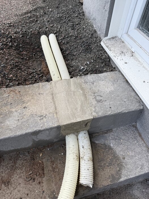 Två dräneringsslangar passerar genom ett gap i en betongtrappa, omgivet av nytt cementarbete. Bakgrund av grus och en vägg syns.