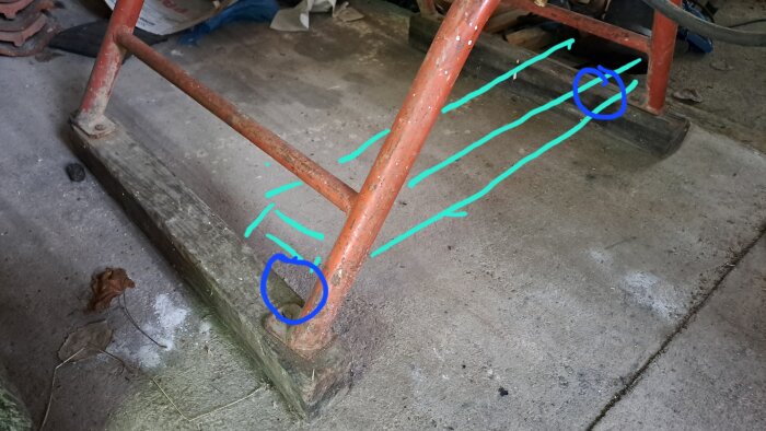 Rött metallstativ på betonggolv med ritade linjer och markeringar som visar byggidé för tillsatta hjul under tvärslåar.
