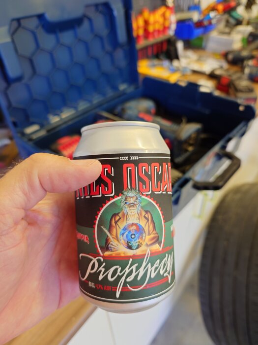 En hand håller en ölburk märkt "Nils Oscar Prophecy" i ett garage med verktyg och däck i bakgrunden.