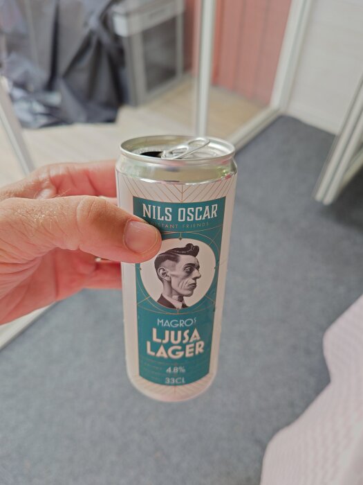 Hand håller en burk Nils Oscar ljusa lager öl, 33 cl, 4,8% alkoholhalt. Bakgrunden visar en suddig uteplats eller balkong.