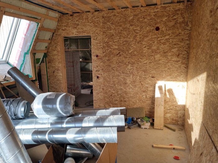 Rum under renovering med OSB-väggar, byggmaterial, ventilationsrör och byggutrustning på golvet.