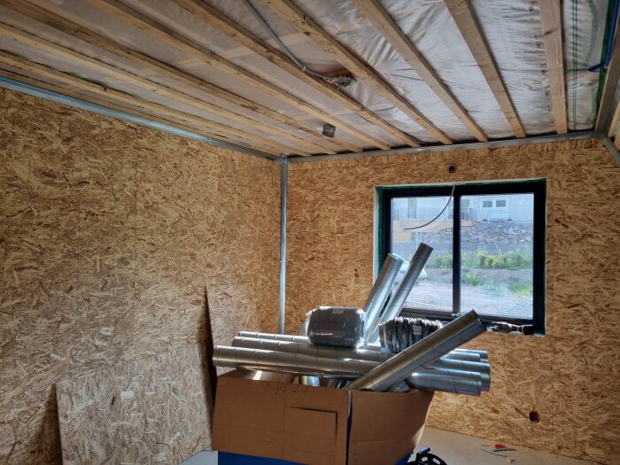 OSB-beklädda väggar och ett delvis isolerat tak, fönster ut mot trädgård. Kartong med ventilationsrör på golvet.
