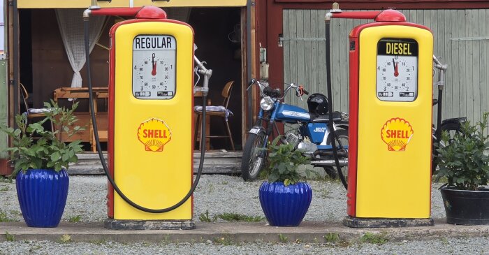 Gula vintage bensinpumpar med Shell-logga, en för vanlig bensin och en för diesel, står bredvid en blå moped och blå blomkrukor.
