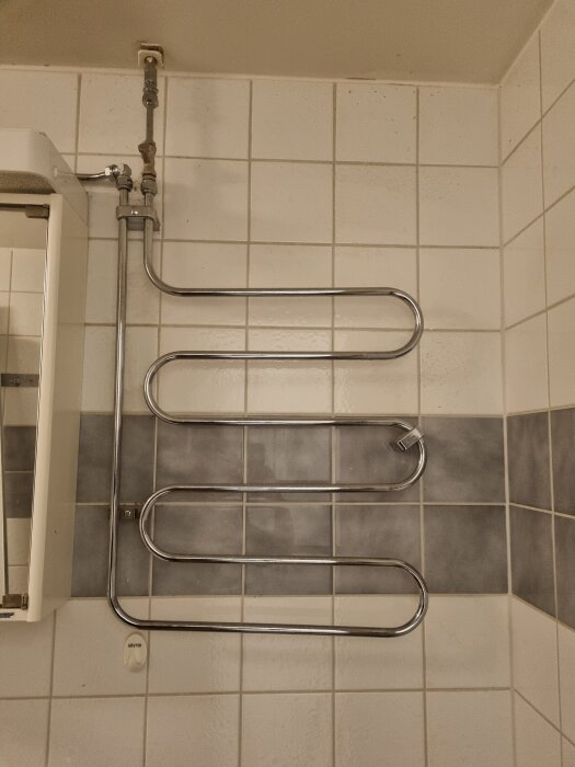 Handdukstork i ett badrum, kopplad till varmvattenrör, monterad på kaklad vägg med vit och grå mosaik.