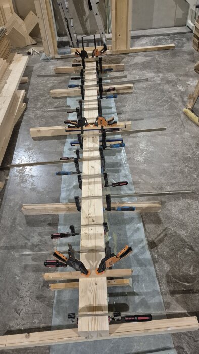 Ett långt trästycke omges av flera tvingar på ett betonggolv, omgiven av byggmaterial.