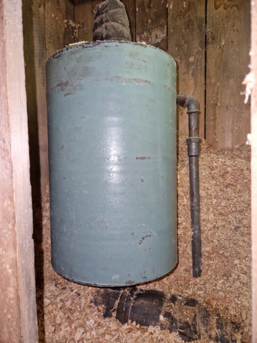 Äldre öppet expansionskärl i plåt med rostfläckar, placerat i ett träbeklätt utrymme med spånströ på golvet. En rörkoppling syns till höger.
