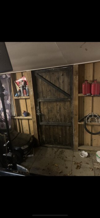 Trädörr och hyllor i ett garage. Hyllorna har objekt som rengöringsprodukter och små dekorationer. Golvet består av marksten med löv.