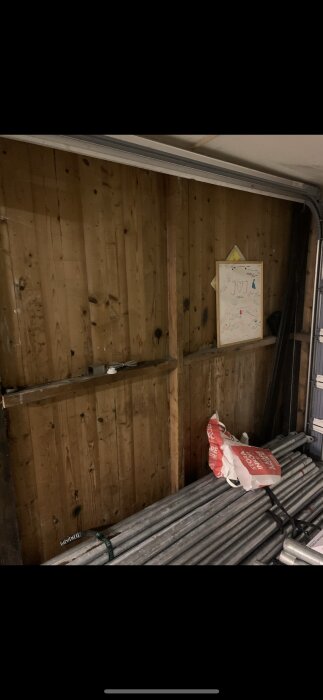 Vägg av träpaneler i ett garage med diverse föremål framför, inklusive metallrör och en papperskasse.