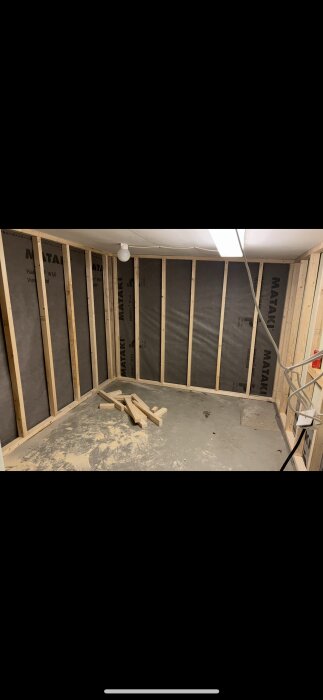 Ett pågående renoveringsprojekt i ett garage med isolerade väggar och synliga reglar, några träbitar ligger på betonggolvet.