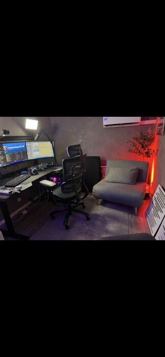 Gamingrum med datorbord, två skärmar och en mikrofon. En kontorsstol står framför, en fåtölj och växt vid sidan, samt röd belysning längs väggen.