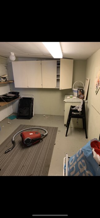 Garage med vita väggskåp, dammsugare på en matta, hopfällbara stolar och ett skåp med duk. En ljusrörslampa i taket.