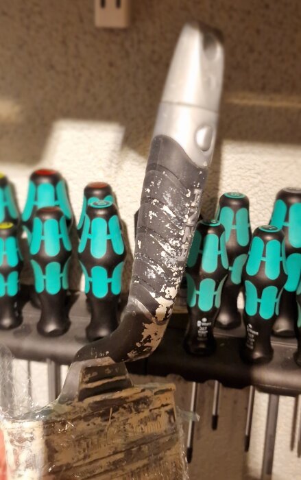 En sliten målarpensel med ett svart och silverfärgat handtag. I bakgrunden syns skruvmejslar med blått och svart handtag.