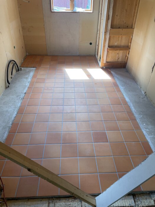 Nyinstallerade Höganäsplattor i rustik rosa på köksgolvet under renovering med träväggar och ett fönster i bakgrunden.