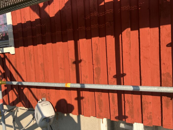 Röd träfasad med skuggor från byggställning och ojämnhet i målningsarbetet. Solen skiner på färgen som ser flagnande ut.