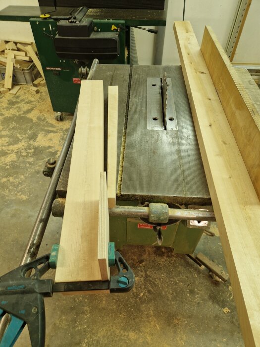 Träbitar och såg på arbetsbänk i snickeriverkstad, förberedelser för att göra ett köksbord av björk med ett grönt snickeribord i bakgrunden.