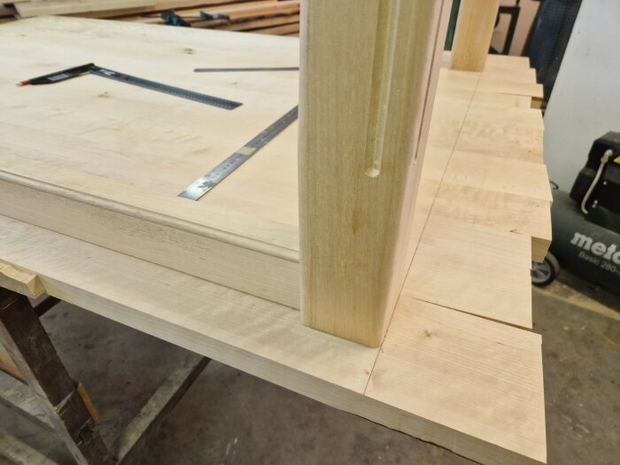 Björkbord under tillverkning i verkstad, med plankor limmade ihop och ett ben monterat. Verktyg, som linjal och vinkelhake, ligger på bordet.