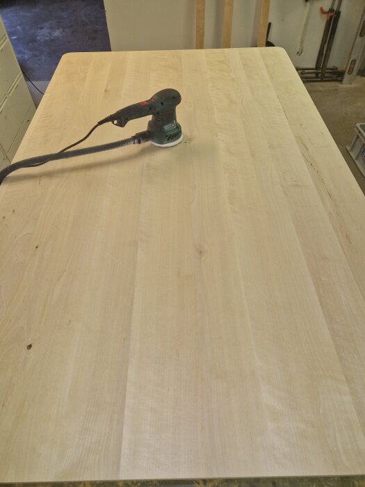 Slipat köksbord av björk med slipmaskin placerad ovanpå. Golvet syns delvis runt bordet i en verkstadsmiljö.