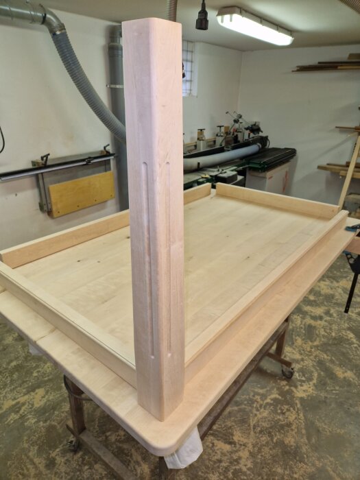 Ett halvfärdigt köksbord i ljust trä står på ett arbetsbord i en verkstad. Bordets ben är avsmalnade och placerade på bordsskivan.