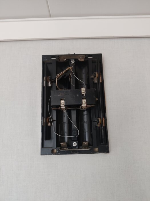 Gammal ringklocka med tom batterihållare och exponerade kablar på vit vägg. Kablar verkar anslutna för extern strömförsörjning.