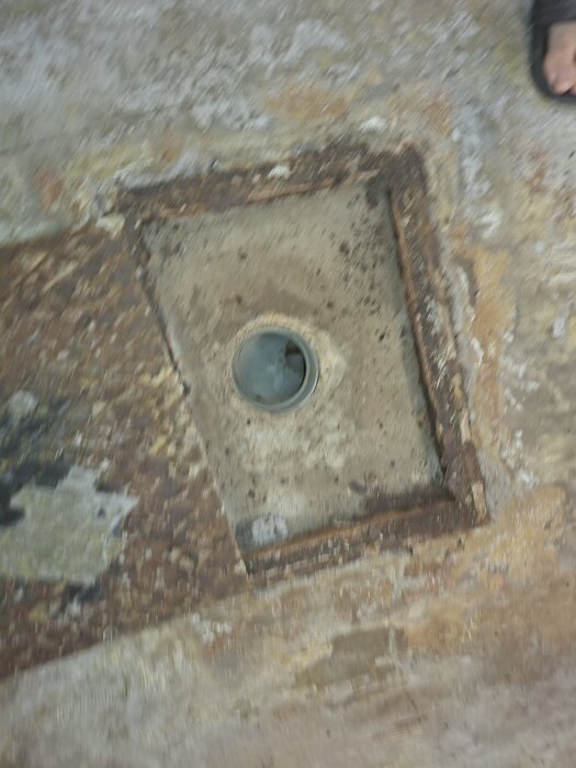 Plastbrunn med metallock försänkt i betonggolv, delvis borttagen plastmatta synlig runt om, avslöjat vid renovering av tvättstuga i enplanshus med källare.