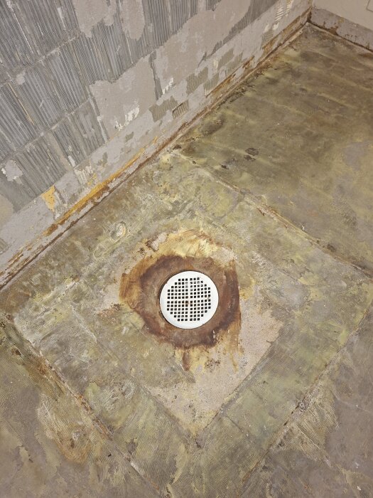 Dolt avlopp i golvet med metallock, delvis nedtaget plastmatta, exponerat betonggolv. Renovationsarbete pågår i tvättstuga i ett enplanshus.