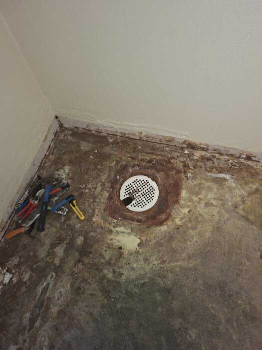 Försänkt plastbrunn med metallock i golvets hörn, omgiven av verktyg; äldre betonggolv i tvättstuga under renovering.