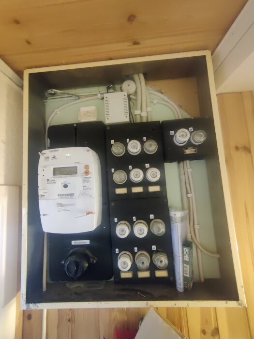 Elcentral med elmätare, säkringar och kopplingsdosor i ett skåp med träpanel i bakgrunden.