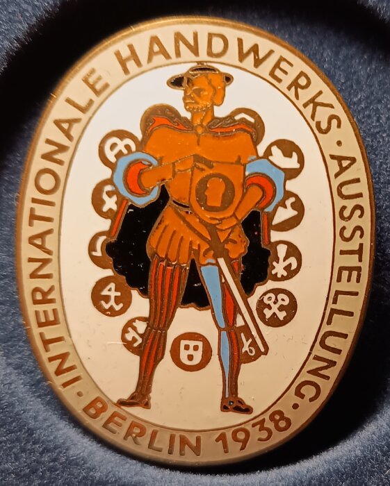 Emaljerat märke från internationell hantverksutställning i Berlin 1938, med figur och text "Internationale Handwerks Ausstellung".