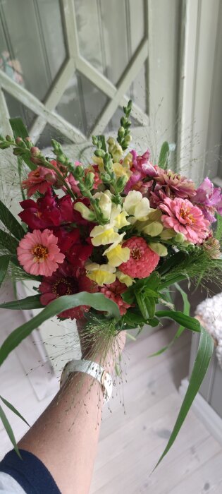 En färgglad bukett med blandade blommor, inklusive rosaröda och gula blommor, hålls av en person med en vit klocka på handleden.