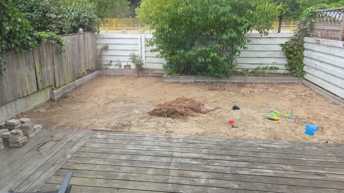 Bakgård med sand, bortplockad marksten, rötter, omgiven av trästaket och buskar. Plastleksaker ligger utspridda på sanden.