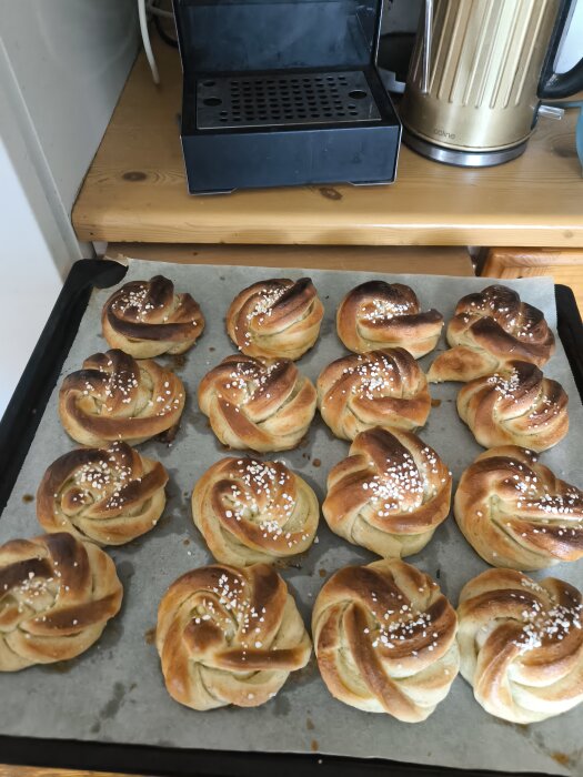 Nygräddade kardemummaknutar med pärlsocker på en bakplåt, bredvid en kaffemaskin och vattenkokare.